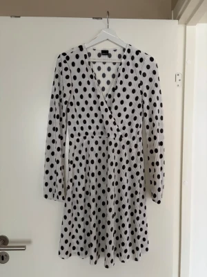 Polka dot klänning  - Supersöt kort vit klänning från Gina med svarta prickar. Omlottmodell med v-ringning och långa ärmar. Tillverkad i ett mjukt, stretchigt tyg som ger en skön passform. Fungerar även om man använder en storlek större 