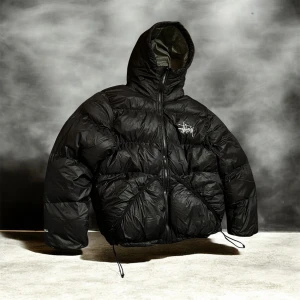 Svart pufferjacka från Stüssy - Svart pufferjacka från Stüssy med huva och dragkedja framtill. Jackan har en oversized passform, synligt Stüssy-tryck på bröstet och snörning vid nederkanten. Materialet är glansigt syntet och jackan är quiltad för extra volym och värme.