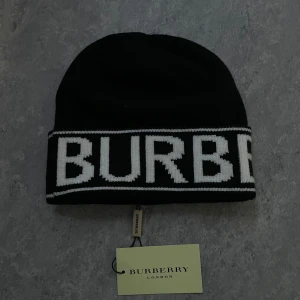 Svart Burberry mössa med logga - Svart mössa från Burberry med stor vit logotyp runt hela kanten. Klassisk och stilren design med ribbad kant och tydlig Burberry-tag. Perfekt accessoar för att lyfta din look med en lyxig touch.