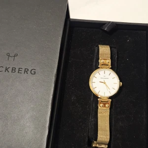 Mockberg guld klocka med mesh-armband - Snygg och stilren klocka från Mockberg med guldigt mesh-armband och vit urtavla. Klockan har minimalistisk design med tunna visare och index, samt ett elegant spänne. Boetten är i rostfritt stål och klockan levereras i originalask med manual. Justerbar band. 