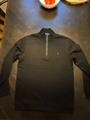 Svart half zip tröja från Polo Ralph Lauren - Svart långärmad tröja från Polo Ralph Lauren med klassisk röd broderad logga på bröstet. Tröjan har en snygg half zip-dragkedja med läderdetalj och stårkrage. Materialet är mjuk bomull som känns skönt mot huden. Perfekt för dig som gillar stilren och enkel design. Mått: Bröstvidd= 53 cm Axlar= 46,5 cm Längd= 70 cm