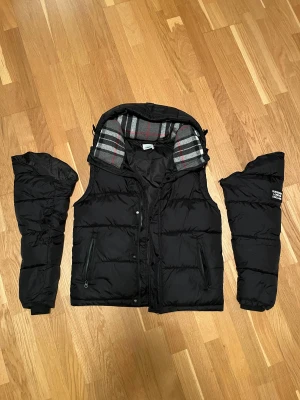 Burberry jacka  - Säljer en svart dunjacka från Burberry London England med avtagbara ärmar så den kan användas som väst. Jackan har rutigt grått innerfoder med röda och svarta detaljer, dragkedja och knappar framtill samt huva. Klassisk Burberry-logga på ärmen. Rikgtigt skön och varm och perfekt inför de kalla tiderna