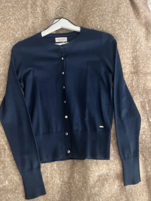 Marinblå kofta från Holly & Whyte - Snygg marinblå kofta från Holly & Whyte by Lindex i storlek S. Den har rund hals, långa ärmar och knappar framtill. Materialet är mjukt och känns som bomull eller en bomullsblandning. Perfekt att slänga på sig när det är lite kyligt ute.