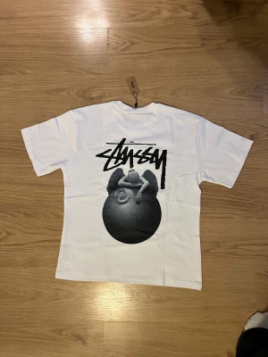 Vit Stüssy t-shirt med tryck, M - Vit t-shirt från Stüssy i bomull med svart tryck på bröstet och stor grafisk print på ryggen med ängel och 8-ball. Klassisk passform och korta ärmar. Aldrig använd