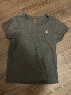 Mörkgrå t-shirt från Polo Ralph Lauren - Snygg mörkgrå t-shirt från Polo Ralph Lauren med klassisk vit broderad logga på bröstet. T-shirten har rund halsringning och korta ärmar. Tillverkad i mjuk bomull för en skön känsla hela dagen.  Stl M men mer som S