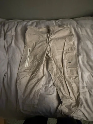 Beige cargopants från Nike, storlek 34 - Säljer ett par beige cargopants från Nike med flera fickor, inklusive en med dragkedja och en med Nike-logga på benet. Byxorna har snörning i midjan och är gjorda i ett lätt bomullsmaterial, perfekta för en chill och sportig stil.