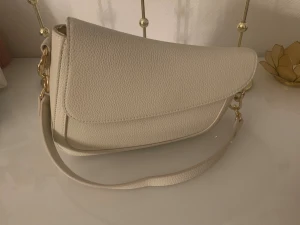 Beige axelväska med gulddetaljer - Snygg beige axelväska med stilrena guldfärgade metalldetaljer. Väskan har en mjukt rundad form och är tillverkad i strukturerat skinnimitation. Perfekt storlek för det viktigaste och har ett lock som stängs med magnet. Enkel och clean design som passar till många outfits.