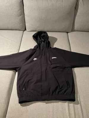 Nike Air Max svart vindjacka med huva - Svart Nike Air Max vindjacka med huva och stora loggor både fram och bak. Jackan har dragkedjor på fickorna, bröstficka med dragkedja och elastisk midja. Tillverkad i syntetmaterial och har en clean, sportig look som passar perfekt till streetwear.