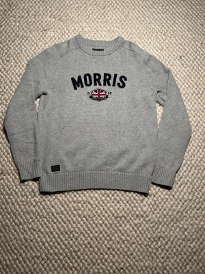Grå stickad tröja Morris S - Snygg grå stickad tröja från Morris i storlek S. Tröjan har broderad Morris-logga och ett Union Jack-emblem framtill. Rund hals och ribbade muddar vid ärmslut och nederkant. Perfekt för dig som gillar klassisk stil med en modern twist.