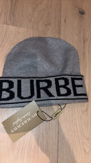 Grå mössa från Burberry - Snygg grå mössa från Burberry med stor logga i svart runt kanten. Tillverkad i mjuk och stickad ull som känns lyxig och varm. Perfekt accessoar för dig som vill ha en stilren och exklusiv look under kalla dagar.