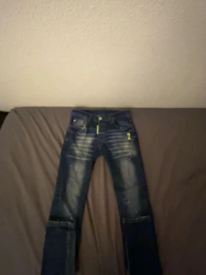 Blå disquared byxor - Snygga blå jeans med slitningar och coola detaljer som gul text på fickan och bakre etikett med 'ICON2'. Jeansen har en skinny passform, slitna knän och ljusa tvättade partier för en trendig look. Perfekt för dig som gillar streetstyle.