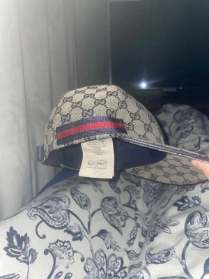 Gucci beige keps med GG-mönster - Beige-grå keps från Gucci med klassiskt GG-monogram över hela kepsen. Kepsen har en justerbar rem baktill, och den har ett rött och 2 blåa ränder på sidan. Materialet är canvas med detaljer i mörkblått tyg. Perfekt för dig som gillar lyxiga accessoarer. Box och allt som följer med medföljer såklart!!