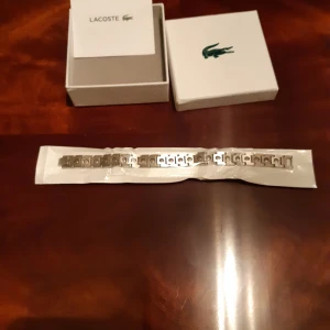 Silverfärgat armband från Lacoste - Snyggt armband i silverfärgad metall från Lacoste. Armbandet har en stilren design med länkade delar och diskreta detaljer. Perfekt accessoar för dig som gillar klassisk och sportig stil. Levereras i originalask med Lacoste-logga.