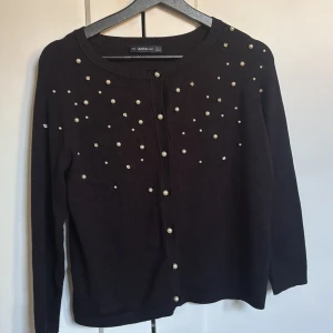 Svart kofta med pärlor från Zara - Svart kofta från Zara med snygga pärldetaljer på framsidan och knappar hela vägen. Stickad och mjuk, perfekt för att lyfta din outfit. Långärmad och med rund halsringning. Passar dig som gillar lite extra detaljer.