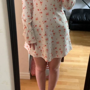 Blommig vit kortklänning  - Supersöt vit kortklänning från NLY One med rosa blommigt mönster. Klänningen har långärmade puffärmar med rynk vid ärmslut och en snygg v-ringning. Materialet är mjukt och lätt, perfekt för varma dagar. Passformen är normal och klänningen har en feminin siluett.