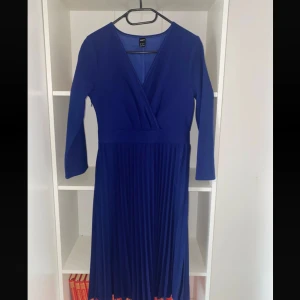 Blå omlottklänning - Snygg blå långklänning med omlottliv och v-ringning. Klänningen har långa ärmar, plisserad kjol och dragkedja i sidan. Perfekt för dig som gillar stilrena och eleganta plagg med en modern twist.