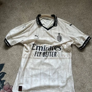 AC Milan x Puma vit fotbollströja - AC Milan x Puma fotbollströja i vitt med subtila mönster och svarta detaljer. Klassisk krage med svart mönster, broderade loggor och tryck från Emirates och Pleasures. Tillverkad i lätt och ventilerande material, perfekt för match eller streetwear.