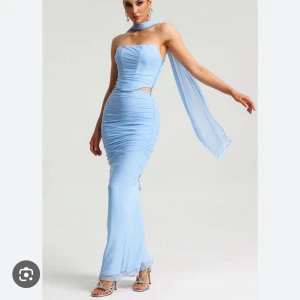 Ljusblå långklänning med scarfdetalj - Säljer en elegant ljusblå långklänning med axelbandslös design och matchande scarf runt halsen. Klänningen har figurnära passform, draperade detaljer och en liten cut-out vid midjan. Perfekt för bal eller fest.