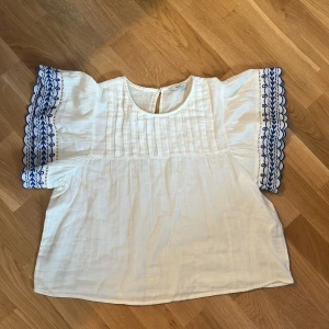 Vit blus med blå brodyr från Pigalle - Supersöt vit blus från Pigalle i luftig linnemix. Vida korta ärmar med blå broderade detaljer och vågig kant. Små veck framtill och knapp i nacken. Perfekt för en fräsch och somrig look!