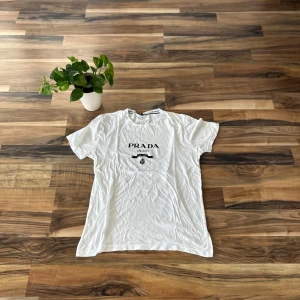 Vit t-shirt från Prada med tryck - Vit t-shirt från Prada med svart logotyptryck på bröstet. Klassisk passform och rund halsringning. Perfekt för dig som gillar stilrena och exklusiva plagg med enkel design.