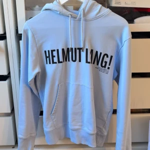 Ljusblå Helmut Lang hoodie - Säljer en ljusblå hoodie från Helmut Lang med stor svart logga framtill. Tröjan har huva med dragsko, magficka och ribbade muddar. Perfekt för dig som gillar stilrena och trendiga plagg med streetkänsla.