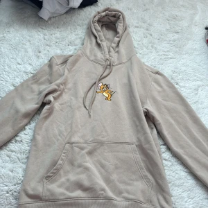 Beige Tom & Jerry hoodie från H&M - Säljer en beige hoodie från H&M med Tom & Jerry-tryck på ryggen och en liten Jerry-brodyr på bröstet. Tröjan har huva med snörning, känguruficka och är i mjuk bomullsblandning. Perfekt för dig som gillar retro vibes och tecknade serier. Använt flera gånger, nypris 370kr.