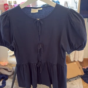 Mörkblå blus med knyt från Gina Tricot - Supersöt mörkblå blus från Gina Tricot med puffiga korta ärmar och tre knyt framtill. Blusen har en lätt volang nedtill och är gjord i mjuk bomull💕pris kan diskuteras men inte under 150kr💕