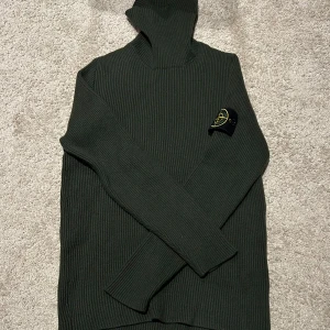 Stone Island grön polotröja i ull - Mörkgrön polotröja från Stone Island med klassisk patch på ärmen. Tröjan är köpt på NK i Göteborg i storlek L men har krympt i tvättmaskinen och sitter nu som en XS. Tröjan har lång ärm, polokrage och är tillverkad i ull. Perfekt för kyliga dagar!