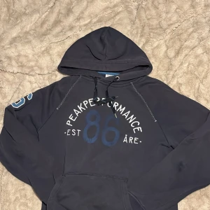 Mörkblå hoodie Peak Performance M - Mörkblå hoodie från Peak Performance med tryck 'Peak Performance 86 Åre' på bröstet och siffran 86 på ärmen. Klassisk huva med snörning och känguruficka framtill. Perfekt för chill dagar eller när du vill ha en sportig look.