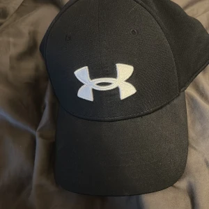 Svart keps från Under Armour L/XL - Svart keps från Under Armour med böjd skärm och stort vitt logotryck framtill. Kepsen är tillverkad i ett mjukt och flexibelt material som sitter skönt på huvudet. Perfekt för sport eller streetwear-stil.