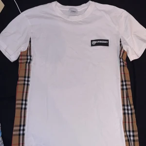 Vit Burberry t-shirt med rutiga sidor - Vit t-shirt från Burberry med klassiska rutiga paneler längs sidorna och diskret Burberry-logga på bröstet. T-shirten har korta ärmar och är gjord i mjuk bomull. Perfekt för dig som vill ha en stilren men ändå unik look.