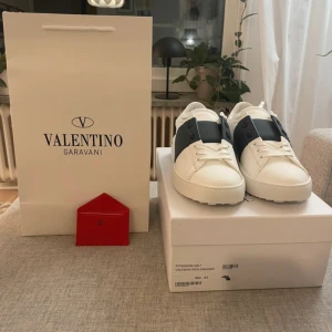 Valentino Garavani Open Sneakers - Snygga vita Valentino Garavani Open sneakers i skinn med bred svart rem över vristen och klassisk snörning. Skorna har en chunky, mönstrad sula och diskret logga på plösen och sulan. Perfekta för dig som vill ha en clean och lyxig look.
