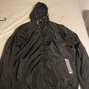 Svart vindjacka från Moncler med huva - Svart vindjacka från Moncler med huva och röda dragsnören. Jackan har dragkedja framtill, två fickor och Moncler-logga på ärmen. Materialet är glansigt och lätt, perfekt för blåsiga dagar. Snygga detaljer med vita och röda accenter vid snörningen.