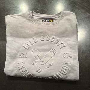 Ljusgrå sweatshirt från Lyle & Scott - Säljer en ljusgrå sweatshirt från Lyle & Scott Junior med stort broderat logotyptryck framtill. Tröjan har rund halsringning och är tillverkad i mjuk bomull, perfekt för en clean och stilren look. Pris kan diskuteras 