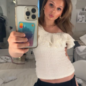 Vit offshoulder topp med volang - Supersöt vit offshoulder topp med volangdetalj längs överdelen. Smockad och figurnära passform som sitter snyggt på kroppen. Perfekt till jeans eller kjol för en trendig look. Tunn och luftig, passar grymt till varmare dagar. Man måste inte ha den offshoulder