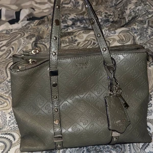 Guess handväska - Snygg grön handväska från Guess med präglat logomönster och silverfärgade nitar på handtagen. Väskan har dubbla dragkedjor, dekorativ nyckel och tag, samt flera fack inuti för smart organisering. Tillverkad i skinnimitation med stilren look. Vet ej om den är äkta