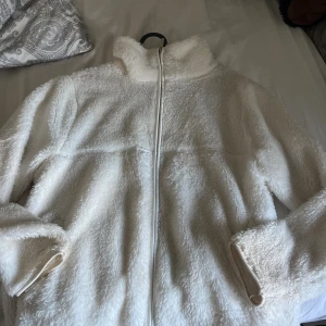 Vit teddyjacka från 157, storlek S - Mysig vit teddyjacka från 157 i storlek S. Jackan har hög krage, hel dragkedja framtill och är tillverkad i fluffigt teddy-material. Perfekt för kyliga dagar när du vill vara både varm och snygg. Enkel och stilren design som är lätt att matcha.