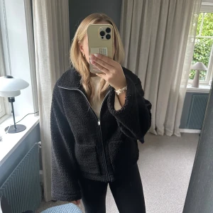 Svart teddyjacka med dragkedja - Mysig svart teddyjacka med hel dragkedja framtill och två stora fickor. Jackan har en oversized passform och är tillverkad i fluffigt teddy-material som håller dig varm. Perfekt för kyliga dagar och enkel att styla till olika outfits.