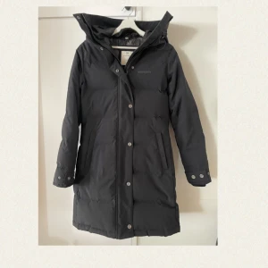 Svart dunjacka i nyskick från Norrsken - Svart varm dunjacka av hög kvalitet från det svenska märket Norrsken. Justerbar huva och öppningsbara tryckknappar nertill på sidorna. Jackan har två fickor framtill och stängs med dragkedja och knappar. Yttermaterial i polyamid, foder i polyester och fyllning av 80% dun och 20% fjäder. Perfekt för kalla dagar. Använd enbart ett fåtal gånger. Nypris 2999kr