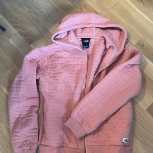 Rosa hoodie från The North Face - Säljer en rosa hoodie från The North Face i storlek M. Tröjan har huva med snörning, dragkedja framtill och ribbade muddar. Materialet är quiltat och känns mjukt och skönt. Perfekt för kyliga dagar när du vill vara både bekväm och snygg.