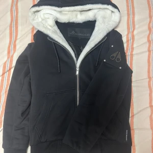 Moos knuckles Bunny - Svart hoodie från Moose Knuckles med vit fluffig fuskpäls i huvan och quiltat innerfoder. Tröjan har dragkedja framtill, snörning i huvan och logga på ärmen. Perfekt för kyliga dagar och riktigt snygg streetstil.