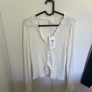 Vit ribbad kofta med volang från Vila - Supersöt vit kofta från Vila med ribbad struktur och volangdetaljer längs v-ringningen, ärmsluten och nederkanten. Långärmad och i mjukt material som känns skönt mot huden. Perfekt att slänga över en topp för en extra touch.