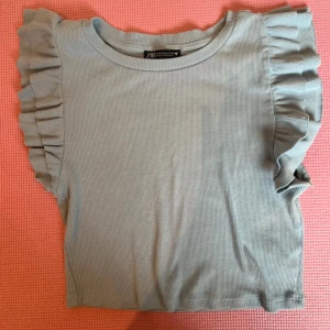 Ljusblå volangtopp från Zara - Supersöt ljusblå topp från Zara med ribbad struktur och volangdetaljer längs axlarna. Toppen är ärmlös och har en rund halsringning. Perfekt för dig som gillar en feminin och trendig stil.