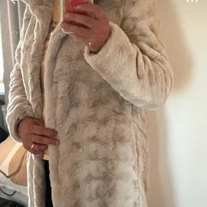 Beige pälsjacka med knappar - Säljer en lång, fluffig beige pälsjacka med snyggt mönstrad yta och knäppning framtill. Jackan har lång ärm och en rak, loose passform som ger en chill vibe. Perfekt för dig som vill ha en varm och trendig jacka med mjuk känsla.