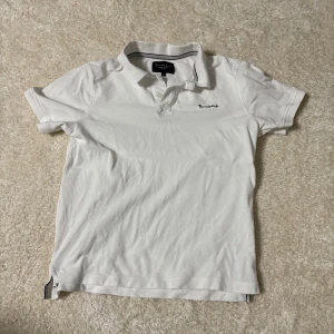 Vit pikétröja från Bondelid, stl S - Klassisk vit pikétröja från Bondelid med liten broderad logga på bröstet. Tröjan har krage, knappar vid halsen och korta ärmar. Tillverkad i mjuk bomull som känns skön mot huden. Perfekt för en clean och stilren look.