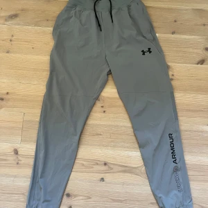 Grå träningsbyxor från Under Armour - Snygga grå träningsbyxor från Under Armour med svart logga på låret och text längs benet. Byxorna har elastisk midja med snörning och är gjorda i ett lätt och smidigt syntetmaterial. Perfekta för dig som gillar sportig stil och vill ha bekväma byxor till gymmet.