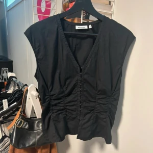 Svart v-ringad topp från Weekday - Svart ärmlös topp från Weekday med v-ringning och knappar framtill. Toppen har rynkade detaljer längs sidorna som ger en snygg form. Perfekt att styla med jeans eller kjol för en cool look.