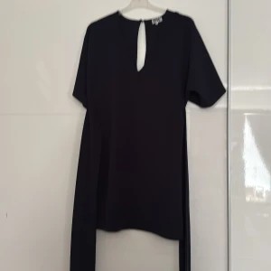 Svart kortklänning från MW - En stilren svart kortklänning från MW med v-ringning och en snygg öppning framtill vid halsen. Klänningen har korta ärmar och är gjord i ett lätt, slätt material som ger en clean look. Perfekt för dig som gillar minimalistisk stil.den har snören som man knyter bak