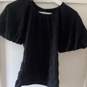 Svart puffärm topp XS - Säljer en svart topp med textur och puffiga korta ärmar. Toppen har en lätt rynkad struktur i tyget och rund halsringning. Perfekt för dig som gillar trendiga siluetter och enkel stil. Helt oanvänd 