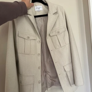Beige overshirt från Josephine H - Stilren beige overshirt från Josephine H med fiskbensmönster, fyra stora fickor framtill och knäppning med knappar. Jackan har klassisk krage och är helfodrad, perfekt för lager-på-lager-stil.